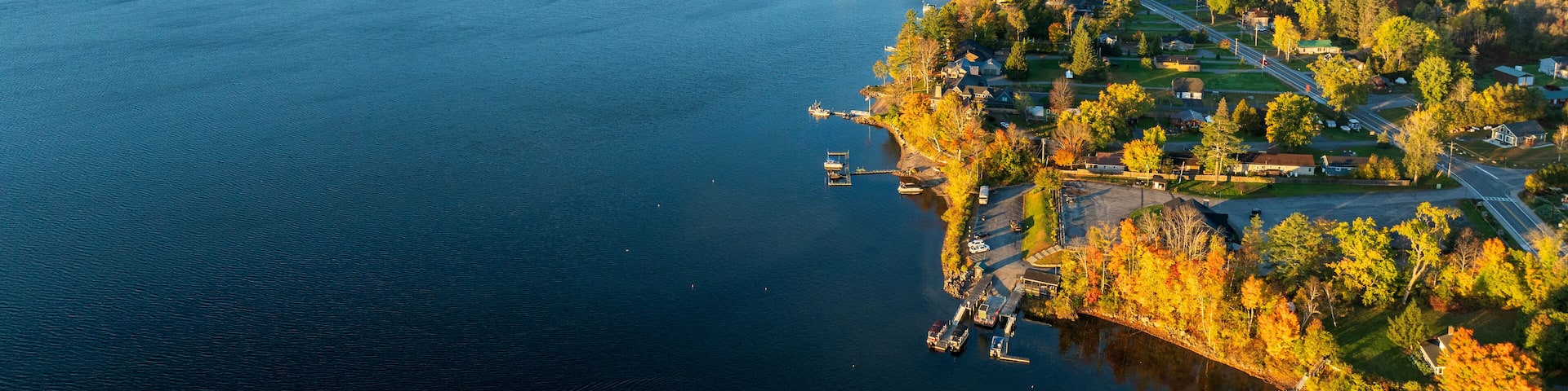 A stunning sunrise over Schroon Lake, NY, with warm light casting a soft glow on the calm water and surrounding autumn-colored forests and mountains.