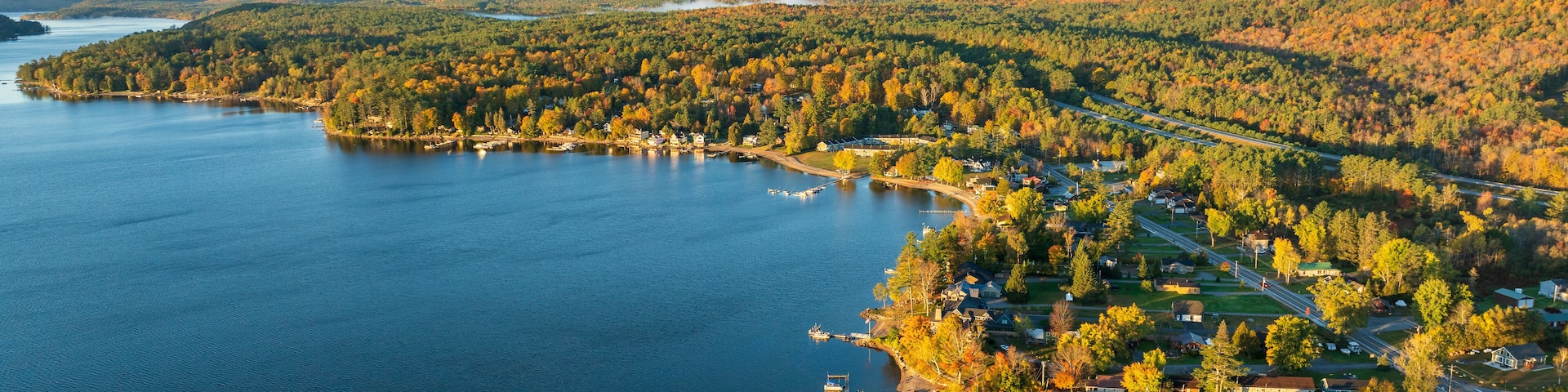 A stunning sunrise over Schroon Lake, NY, with warm light casting a soft glow on the calm water and surrounding autumn-colored forests and mountains.
