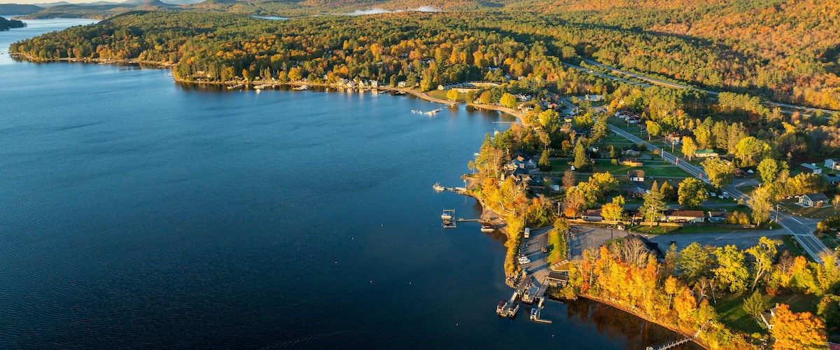 A stunning sunrise over Schroon Lake, NY, with warm light casting a soft glow on the calm water and surrounding autumn-colored forests and mountains.