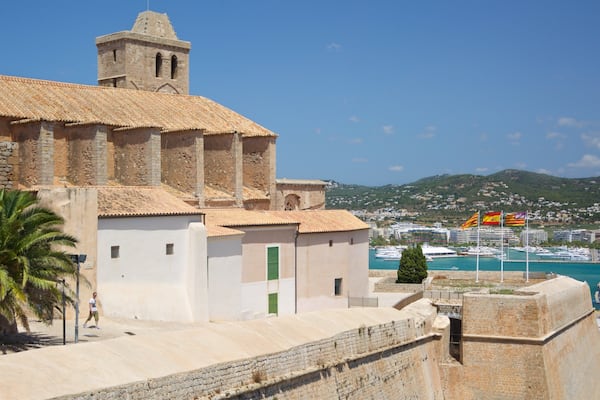 A coastal town with a historic church, palm trees, and a view of the sea with boats and flags.