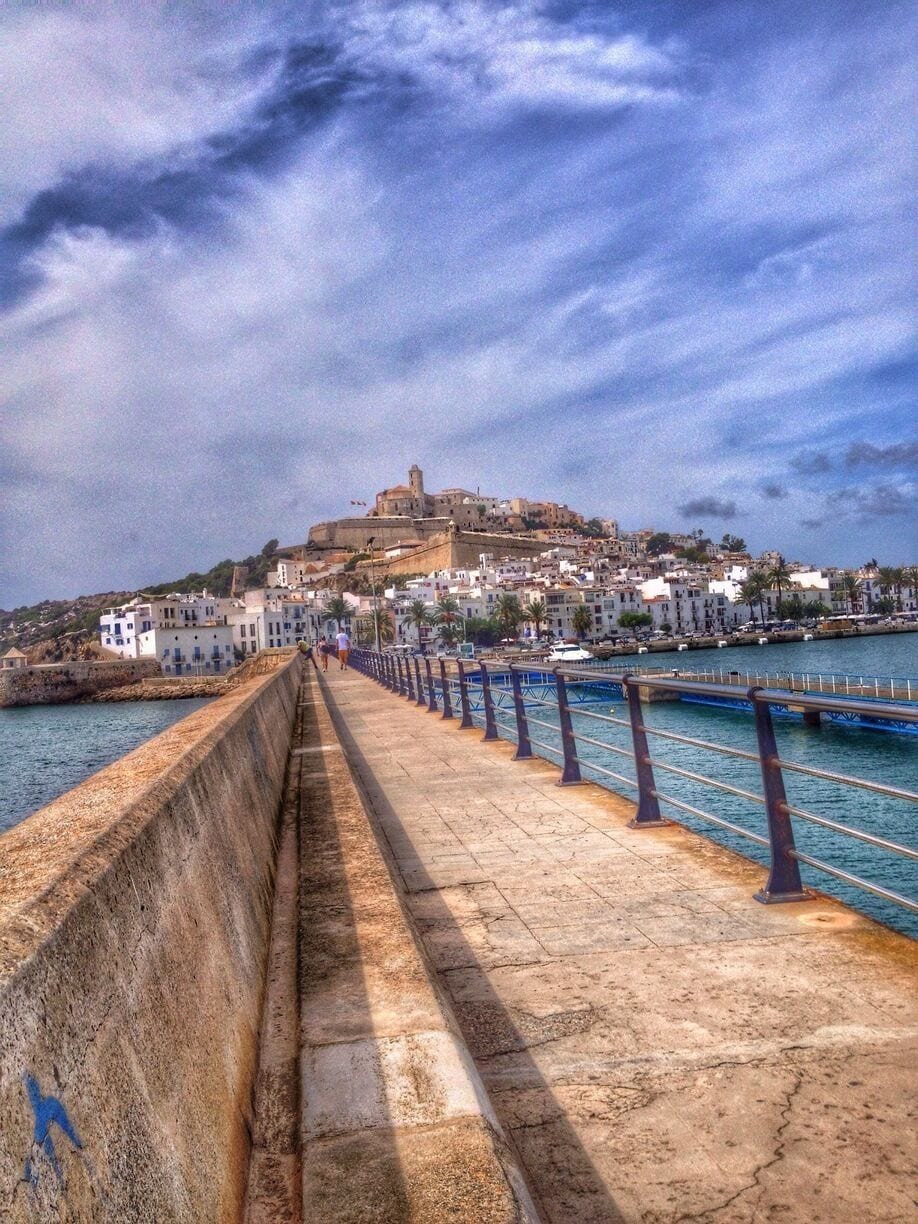 Un paseo marítimo con sendero de piedra, una fortaleza en la colina a lo lejos y un mar tranquilo.