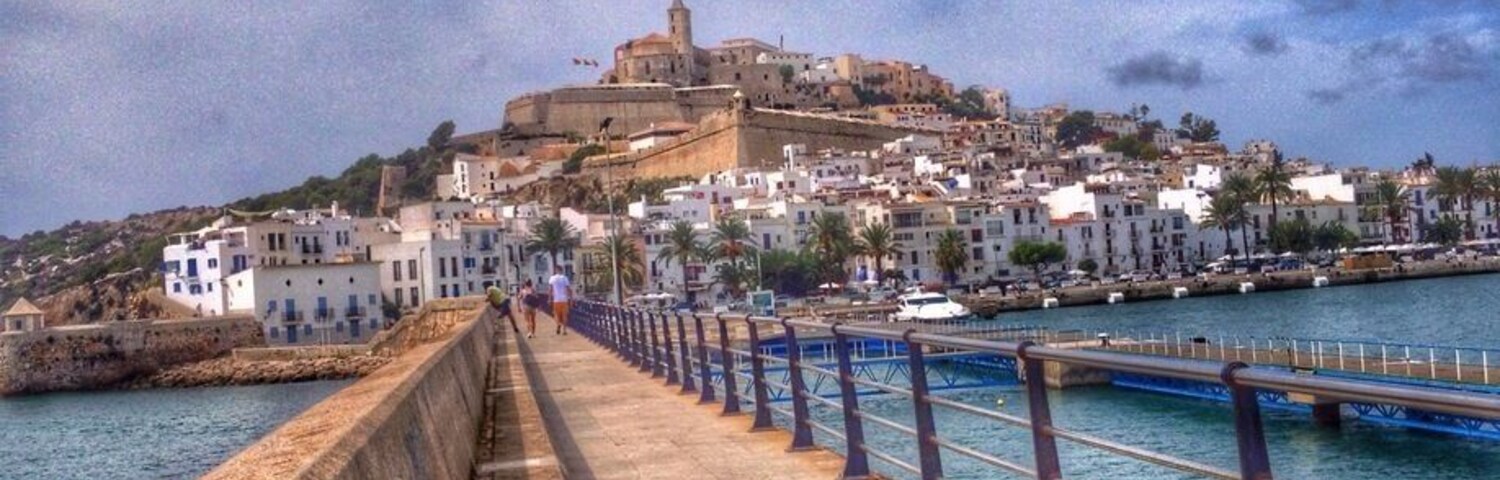Un paseo marítimo con sendero de piedra, una fortaleza en la colina a lo lejos y un mar tranquilo.