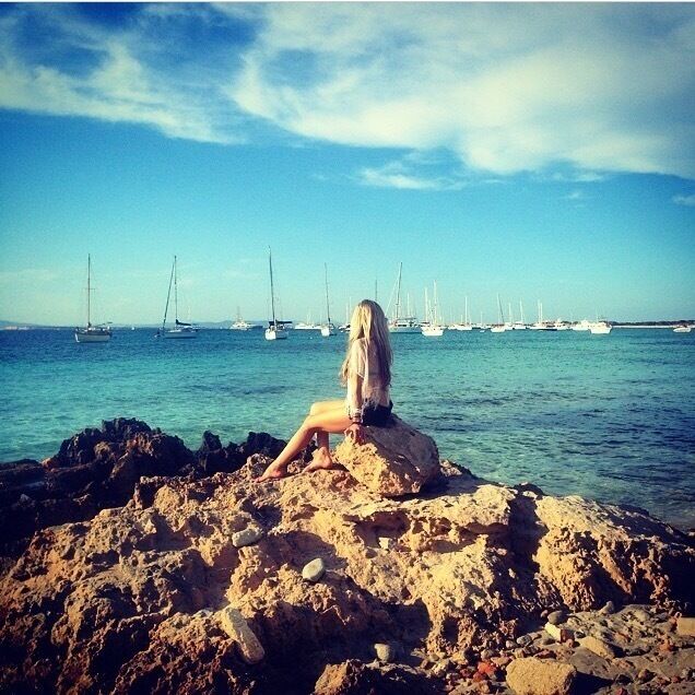 A rocky beach with a person sitting on a rock, overlooking a calm sea with several sailboats.