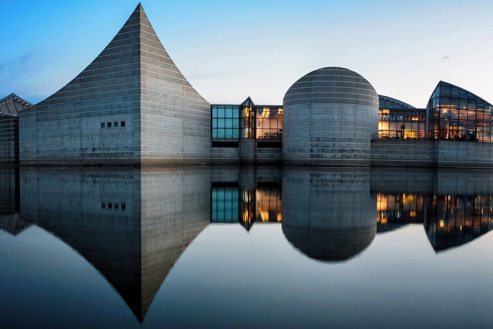 Ein moderner architektonischer Komplex mit einem pyramidenförmigen Gebäude, einer runden Struktur und einem gebogenen Glasgebäude, das sich im Wasser spiegelt.