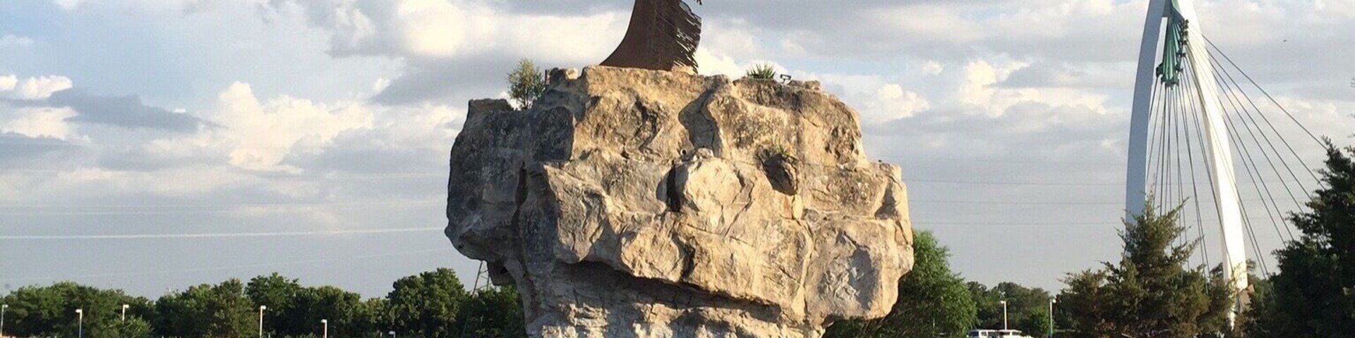 Standing 44 feet atop a large Boulder at the confluence of the Arkansas and Little Arkansas Rivers is the Keeper of the Plains. Designed in 1974 by Comanche-Kiowa artist Blackbear Bosin, the sculpture has since become a symbol of the city of Wichita. At night large fire pits located around the keeper are lit and illuminates the warrior in a fiery blaze.