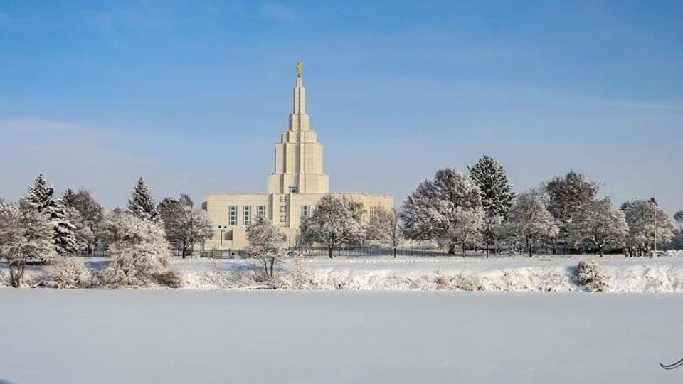 A snow-covered landscape with a prominent building featuring a tall spire, surrounded by trees.