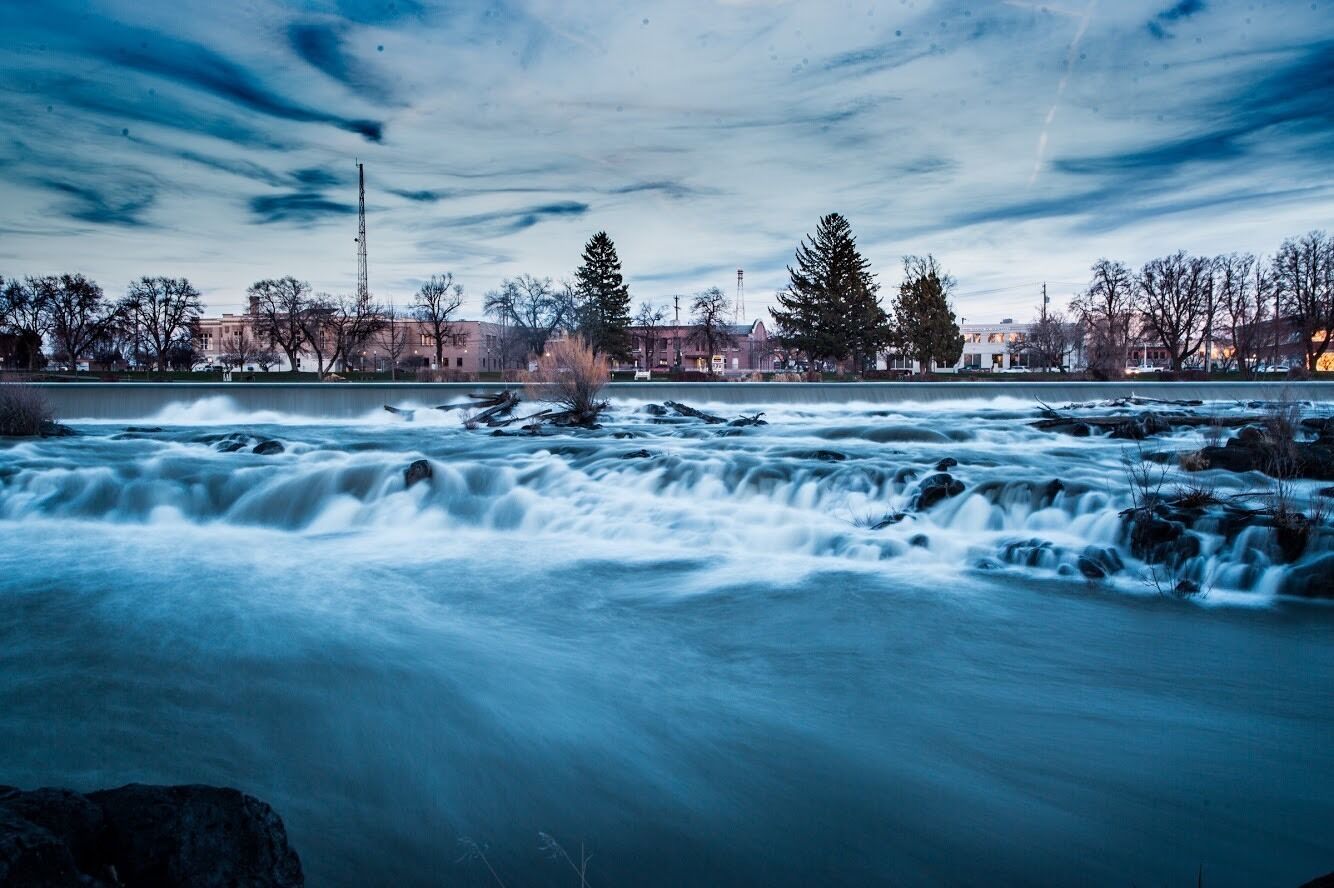 A river with a fast current, surrounded by trees and buildings in the background.