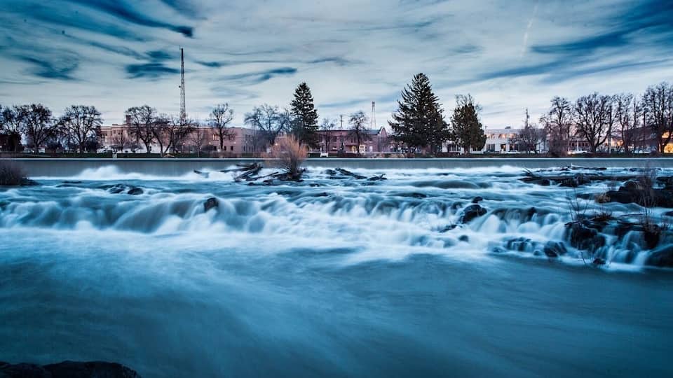 A river with a fast current, surrounded by trees and buildings in the background.