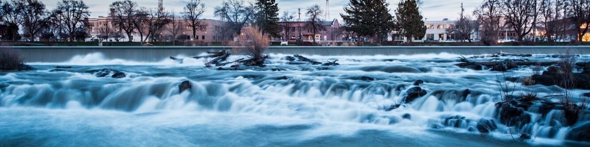 Un río de fuerte corriente, rodeado de árboles y edificios al fondo.