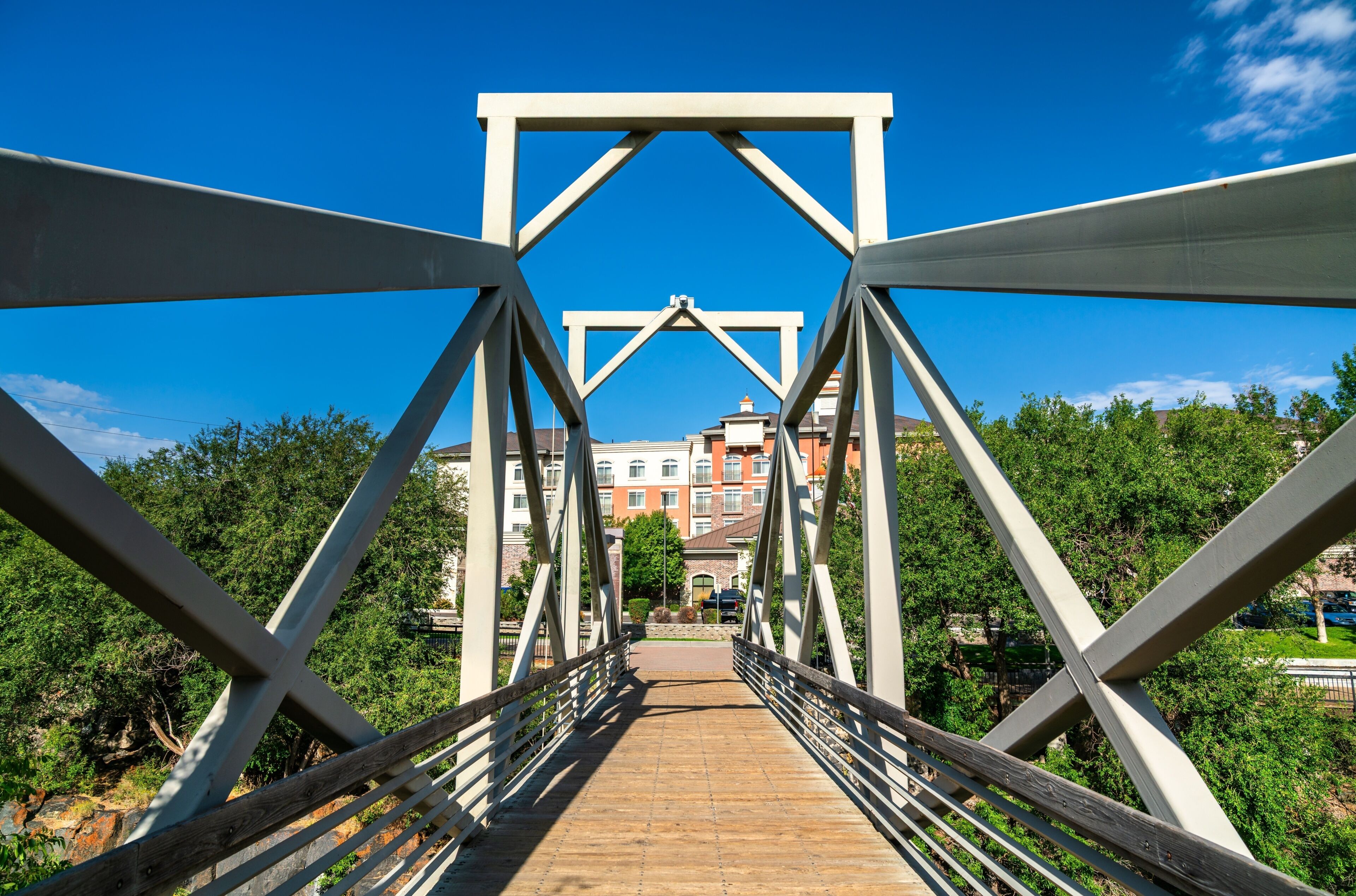 A pedestrian bridge with a metal framework and wooden walkway, leading to a building with a red facade.