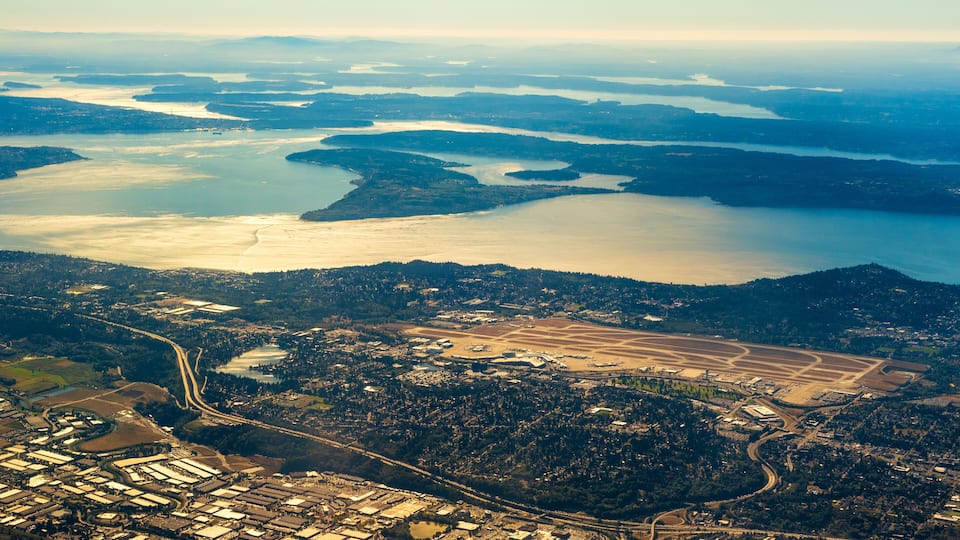 An aerial view of a coastal city with a large body of water, industrial areas, and dense forested regions.