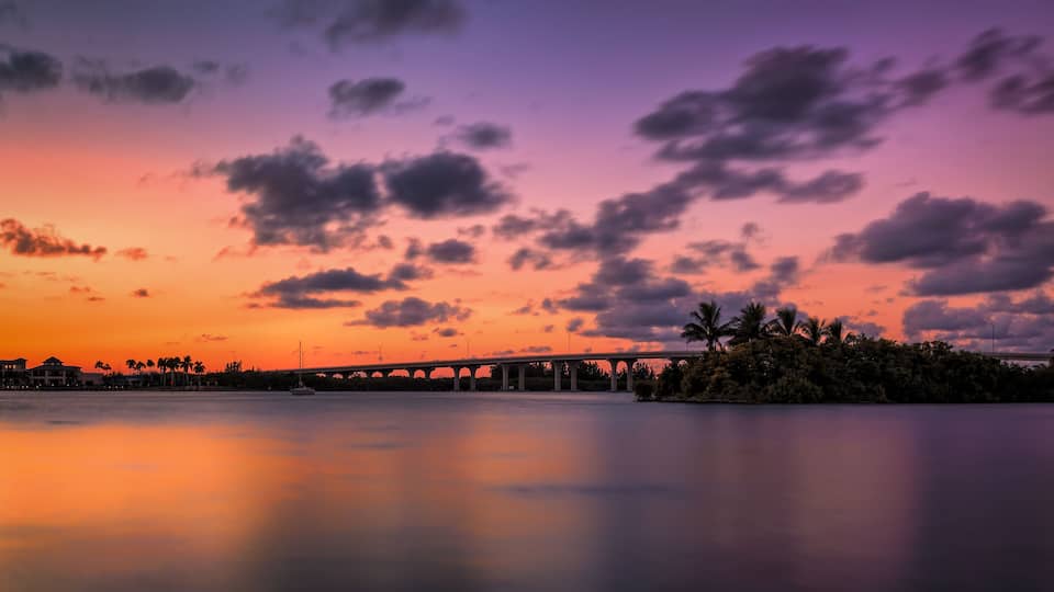 A sunset over a bridge with palm trees and a calm body of water.