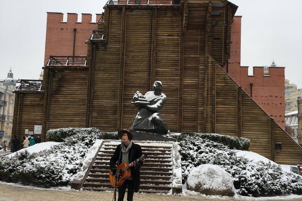 Un musicien joue de la guitare devant un bâtiment historique, sur lequel on aperçoit une statue et un escalier.