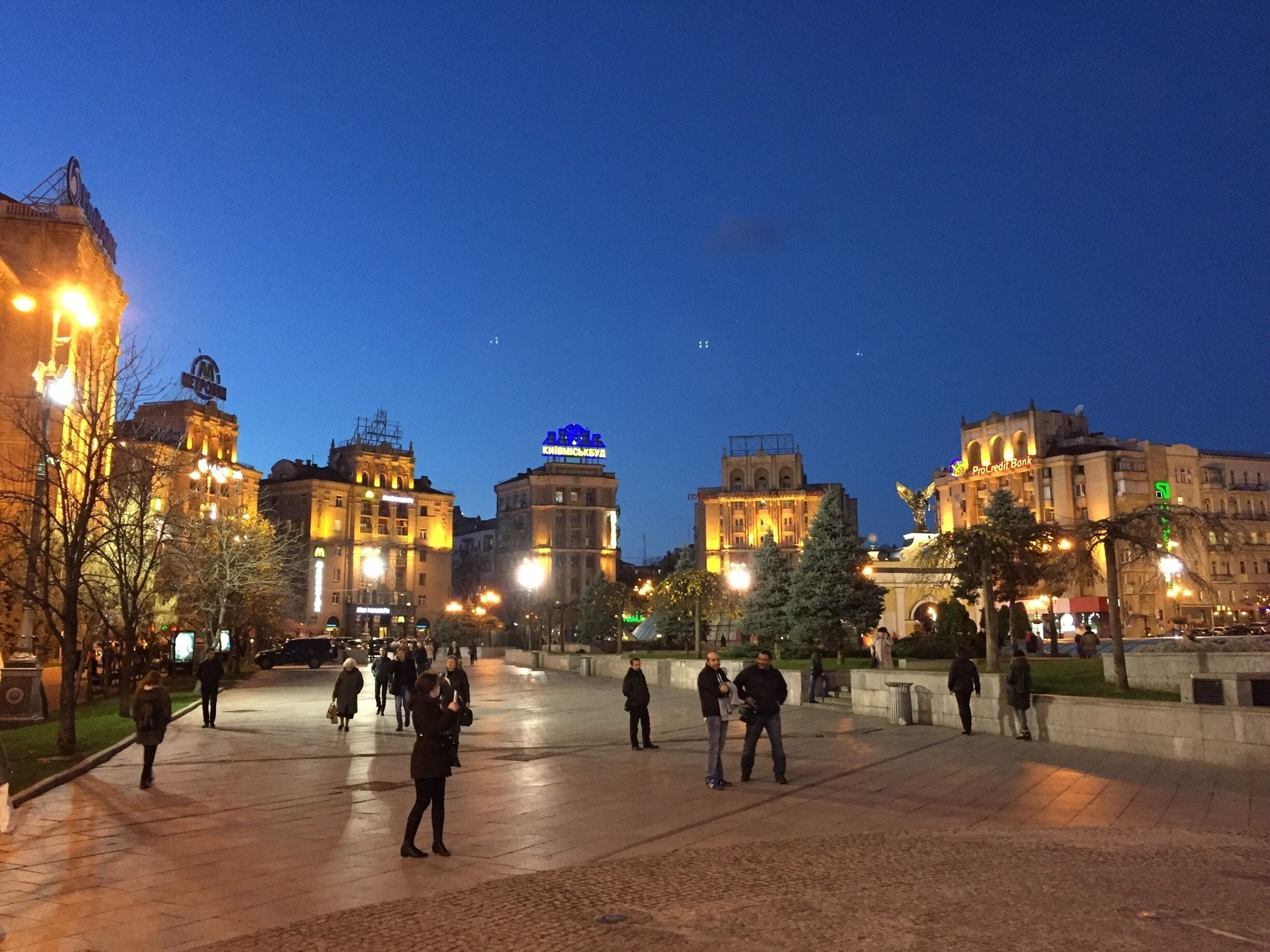 Una plaza urbana de noche con edificios iluminados y gente caminando.