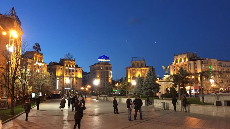 Una plaza urbana de noche con edificios iluminados y gente caminando.