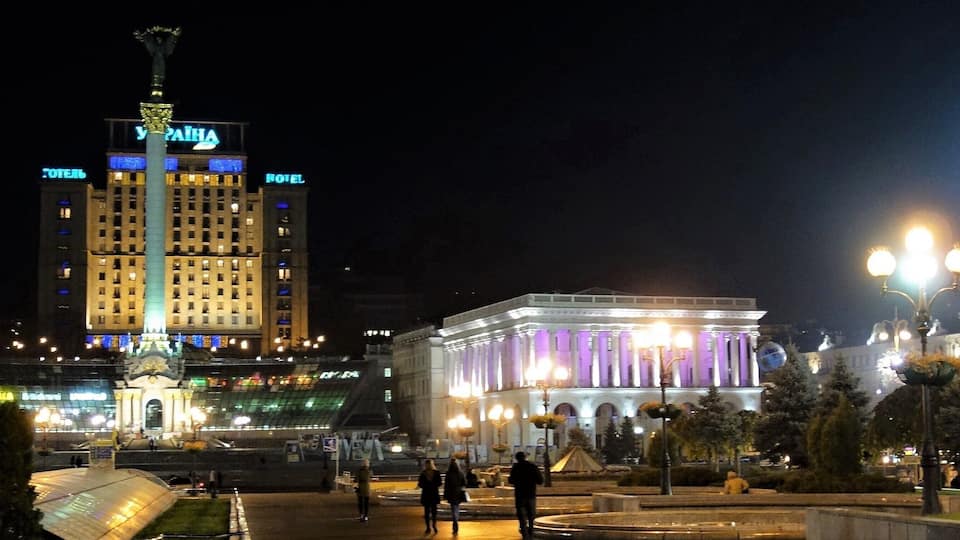 A city square at night with illuminated buildings, including a hotel and a building with purple lighting.