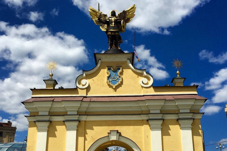 A yellow archway with a golden statue of an angel holding a sword, a sun emblem, and a shield with a blue and white design.