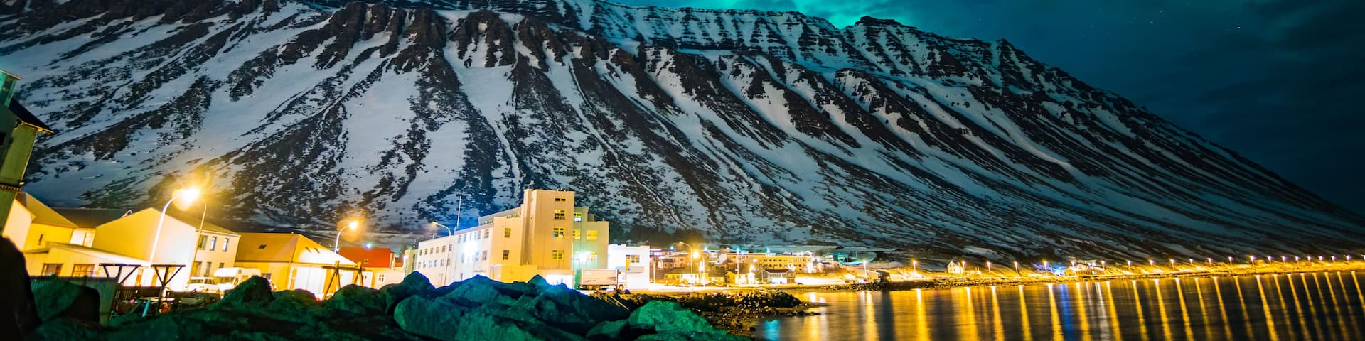 A snow-covered mountain with a lit-up town at its base and the aurora borealis in the sky.