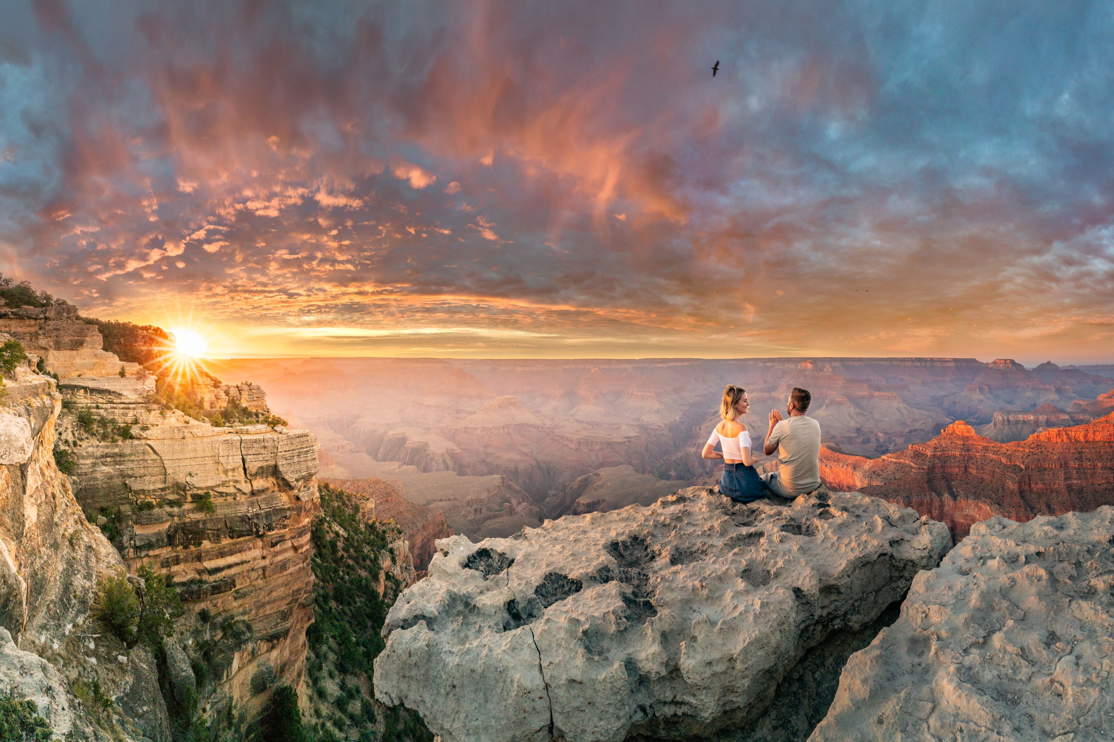Una pareja sentada sobre un saliente rocoso con vistas a un cañón y un atardecer de fondo.