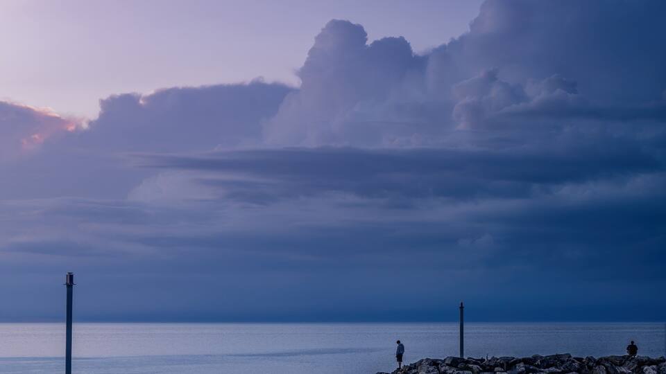 A calm sea at dusk with a rocky breakwater and a lighthouse in the distance.