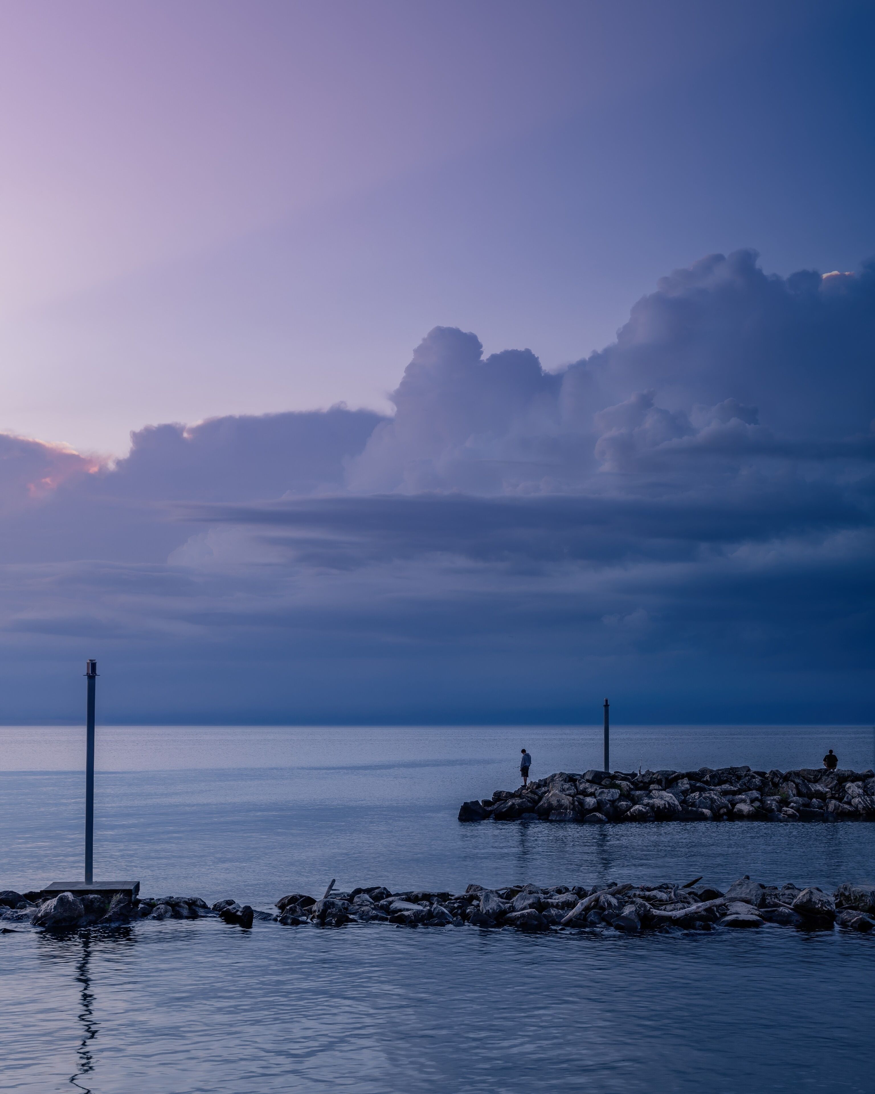 A calm sea at dusk with a rocky breakwater and a lighthouse in the distance.