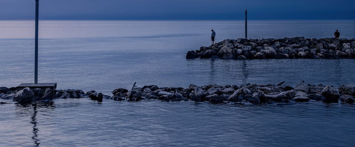 A calm sea at dusk with a rocky breakwater and a lighthouse in the distance.