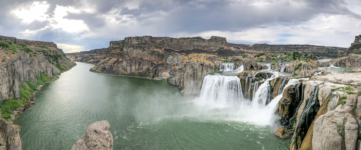 A river canyon with multiple waterfalls, rocky cliffs, and a distant building.