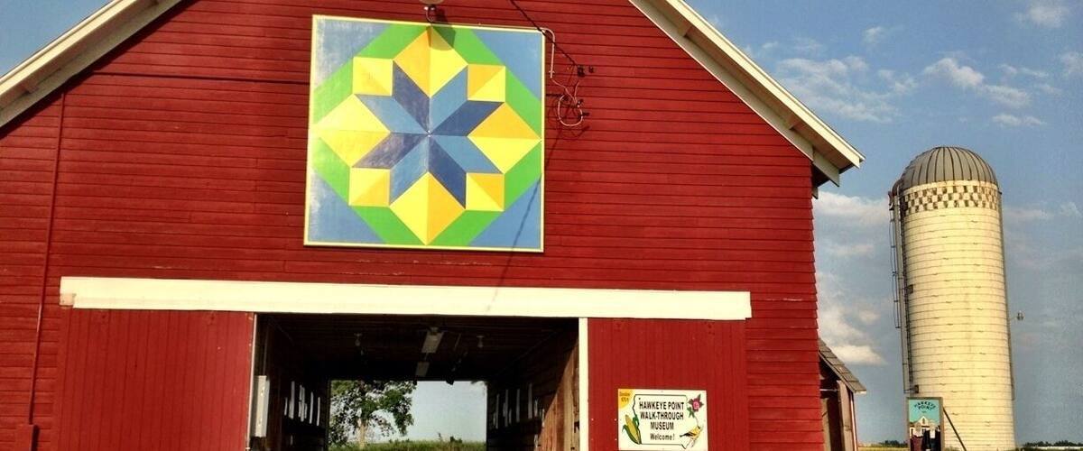 A red barn with a colorful geometric quilt hanging on the front, a picnic table in front, and a silo in the background.