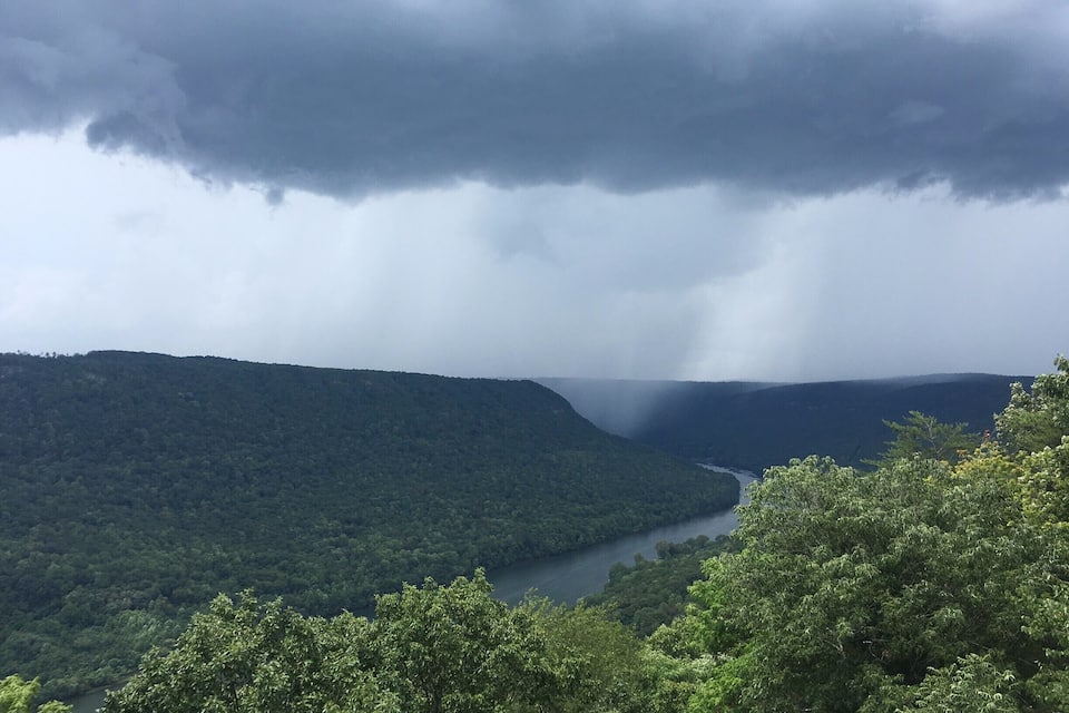 Ein Fluss schlängelt sich durch ein dicht bewaldetes Tal mit steilen Hügeln und einem bewölkten Himmel.