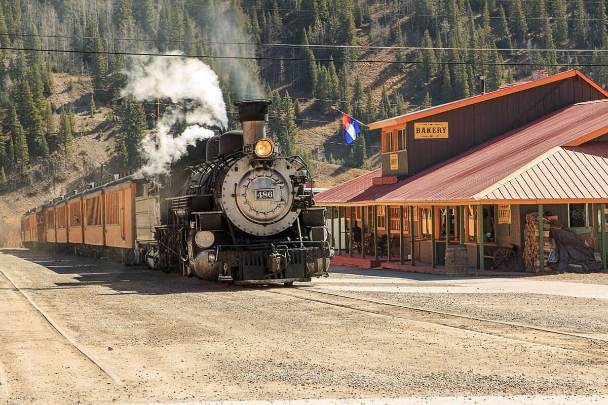 A steam locomotive numbered 456 on tracks, a bakery building, and a mountainous forested backdrop.