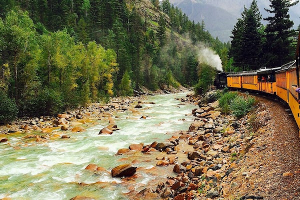 A train traveling along a river surrounded by rocky terrain and dense forest.