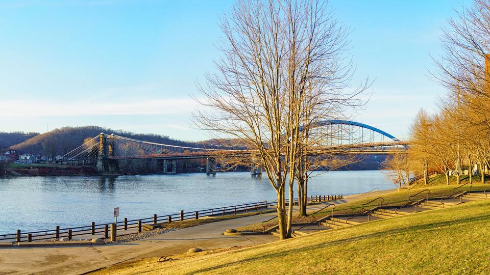 A suspension bridge over a river, a park with a walking path, and leafless trees.