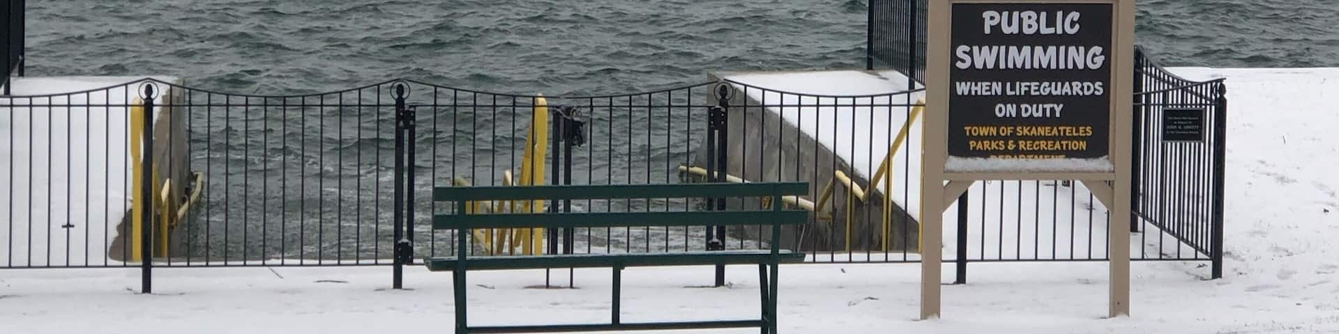 A snow-covered bench, a sign indicating public swimming when lifeguards are on duty, and a view of a body of water with distant land.