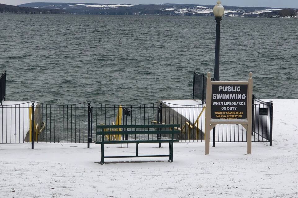 A snow-covered bench, a sign indicating public swimming when lifeguards are on duty, and a view of a body of water with distant land.
