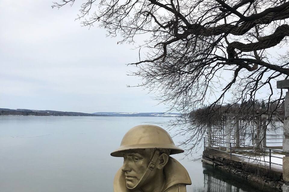 A bust sculpture of a person wearing a helmet and uniform, placed on a stone pedestal by a lake with a dock in the background.