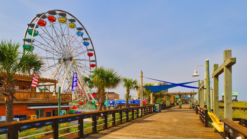 A wooden boardwalk with a Ferris wheel, palm trees, and a beach in the background.
