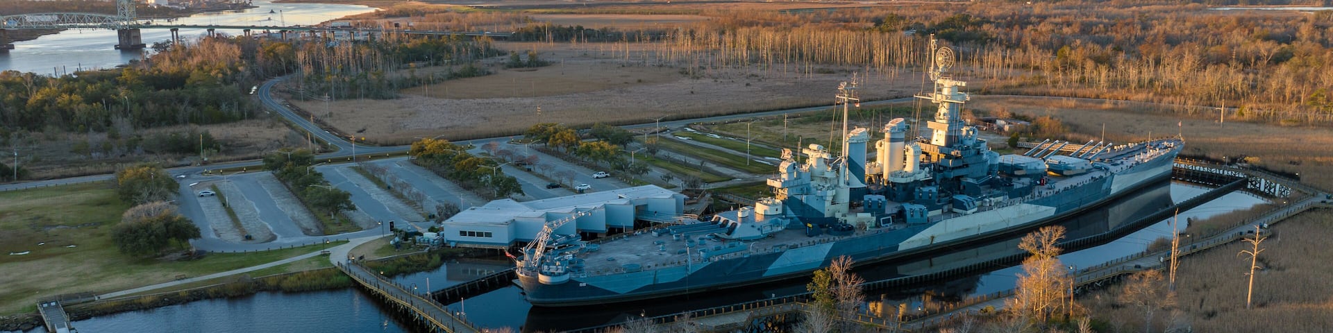 Battleship at sunrise in Wilmington, North Carolina, with the Cape Fear River flowing on the left and the Cape Fear Memorial Bridge in the distance, set against a backdrop of the awakening city.