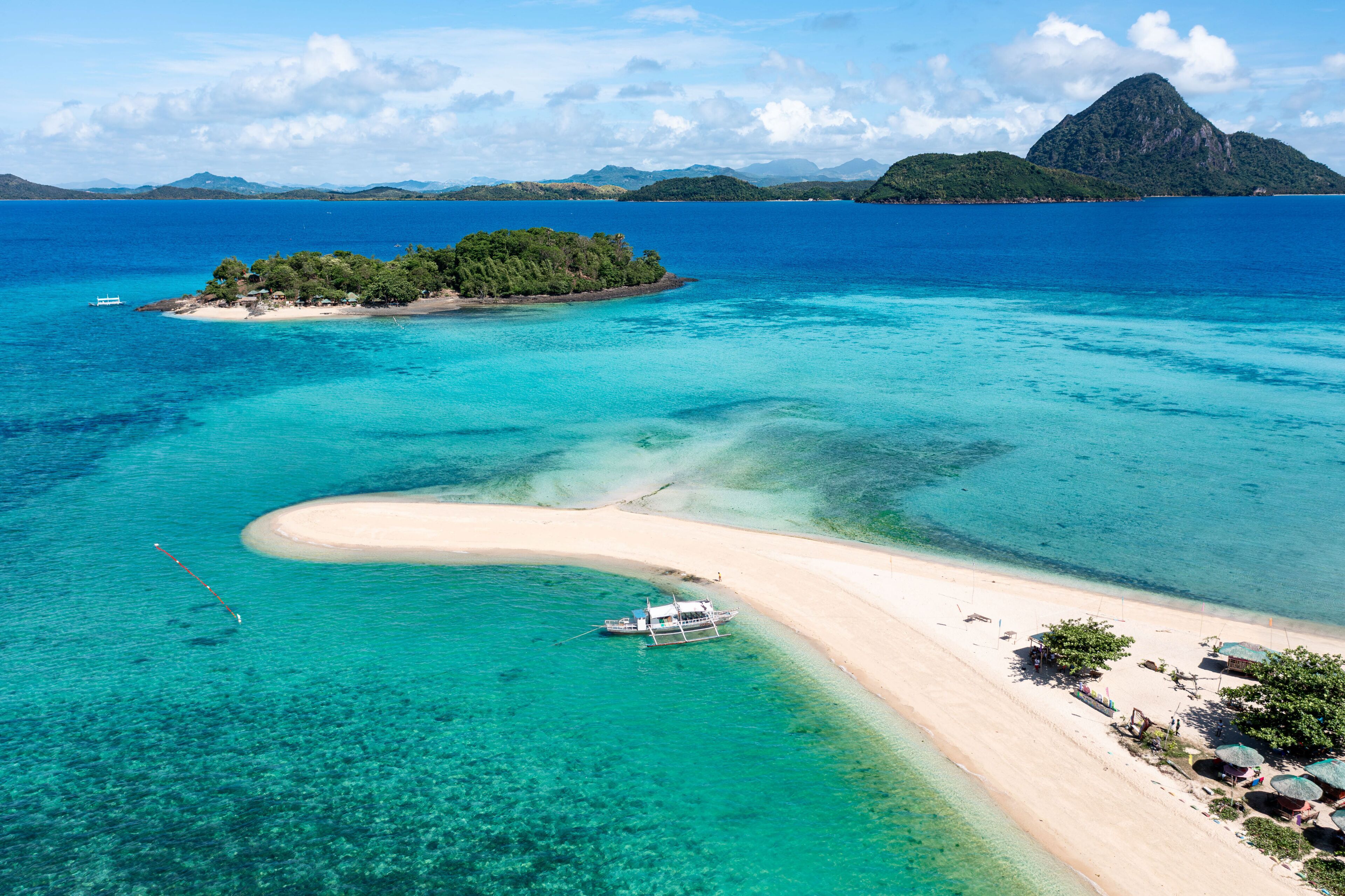 Aerial view of a pristine sandbar stretching into turquoise waters, connecting to a lush island paradise under a bright sky, Concepcion, Western Visayas, Philippines.