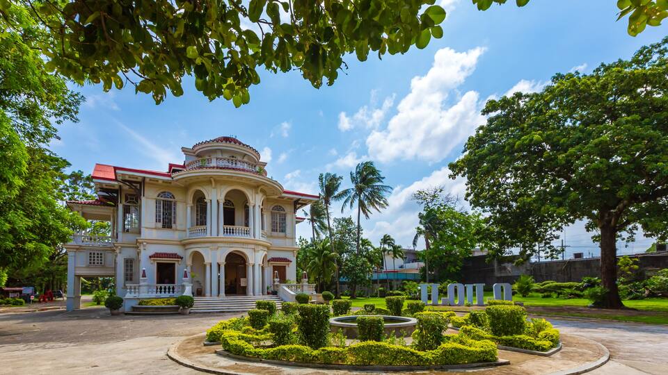 A grand colonial-style building with a red roof, surrounded by well-manicured gardens and palm trees.