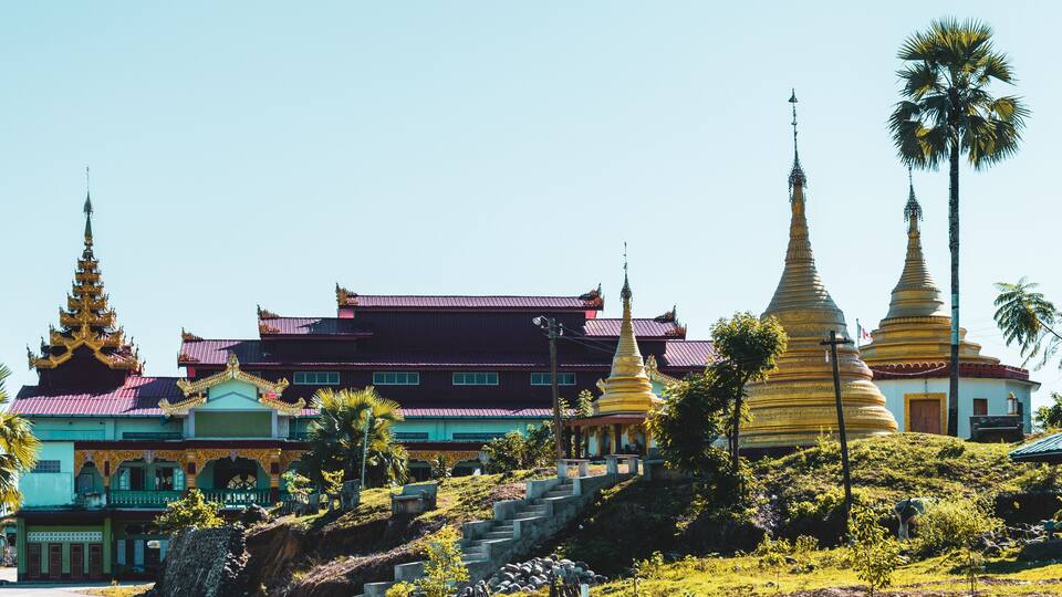 A traditional temple complex with ornate golden spires, a multi-tiered roof, and a lush green landscape.