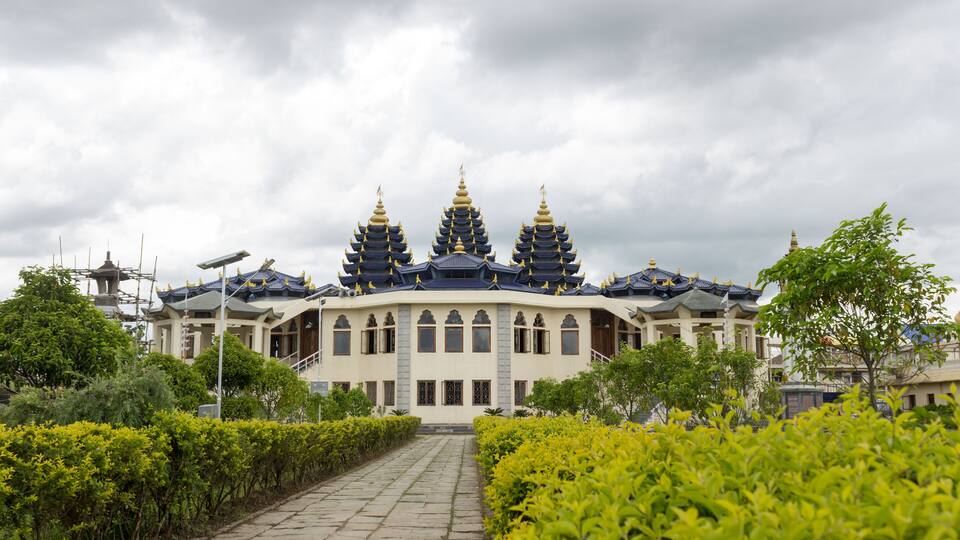 A traditional temple with three pagoda-style roofs, surrounded by well-maintained greenery and a stone pathway leading to the entrance.