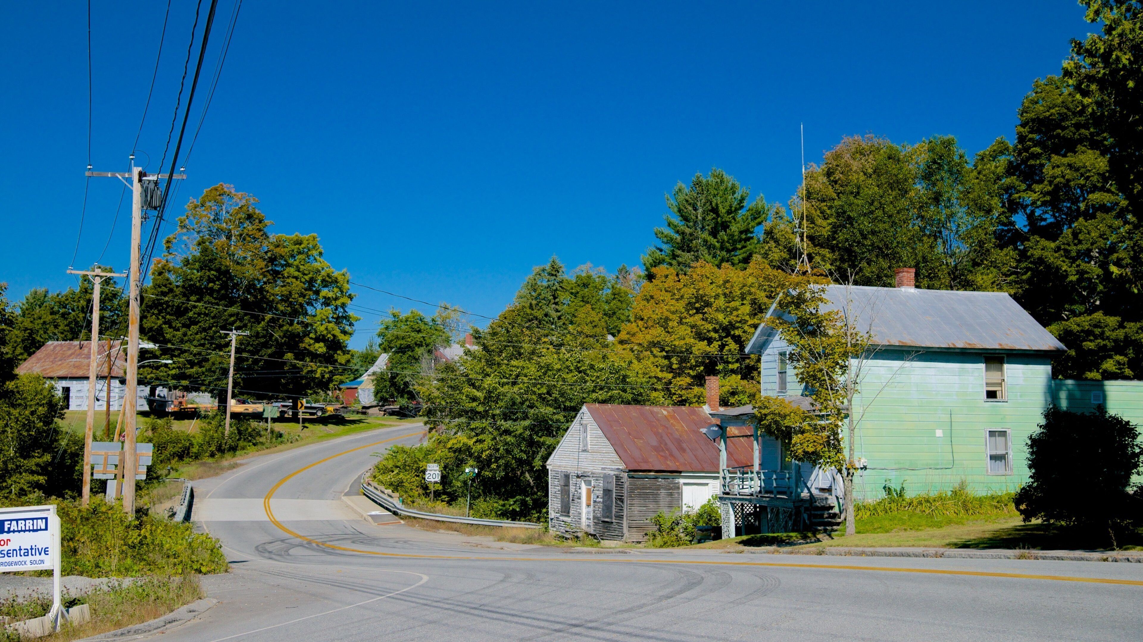 A rural road curving through a small town with old wooden houses, a sign for FARRIN or Bridgework Solon, and lush green trees.