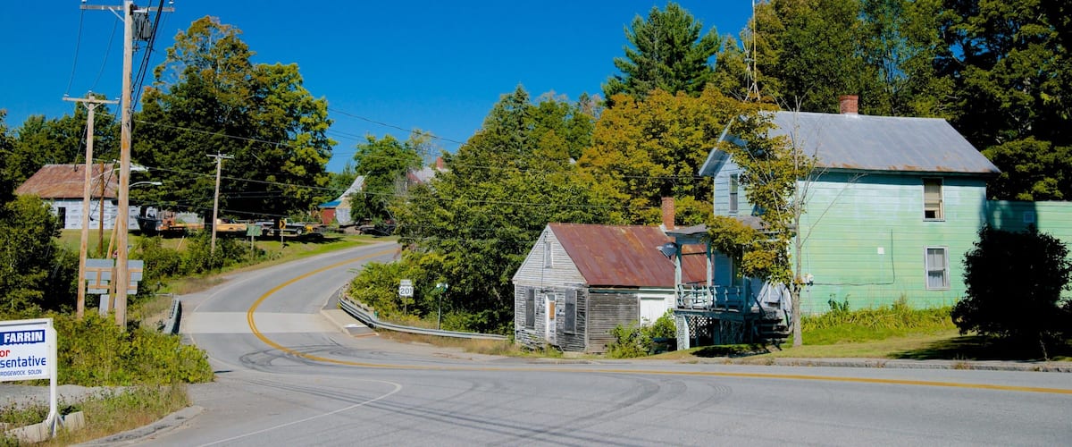 A rural road curving through a small town with old wooden houses, a sign for FARRIN or Bridgework Solon, and lush green trees.