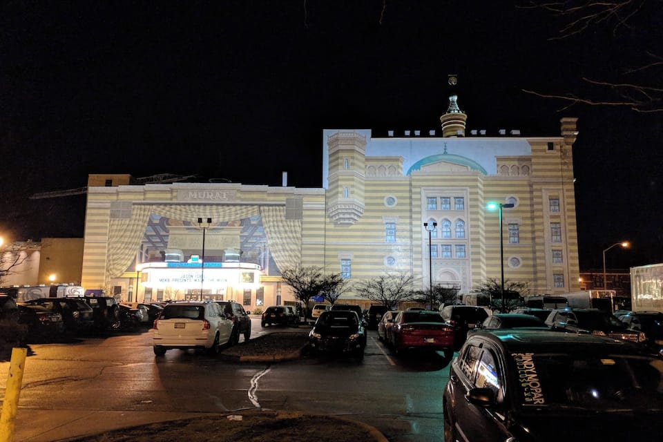 Un edifici gran i il·luminat amb un mural a l'entrada, envoltat de cotxes estacionats.