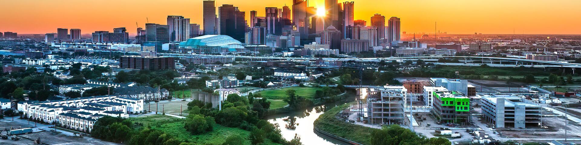 A city skyline at sunset with a river in the foreground and a stadium in the center.
