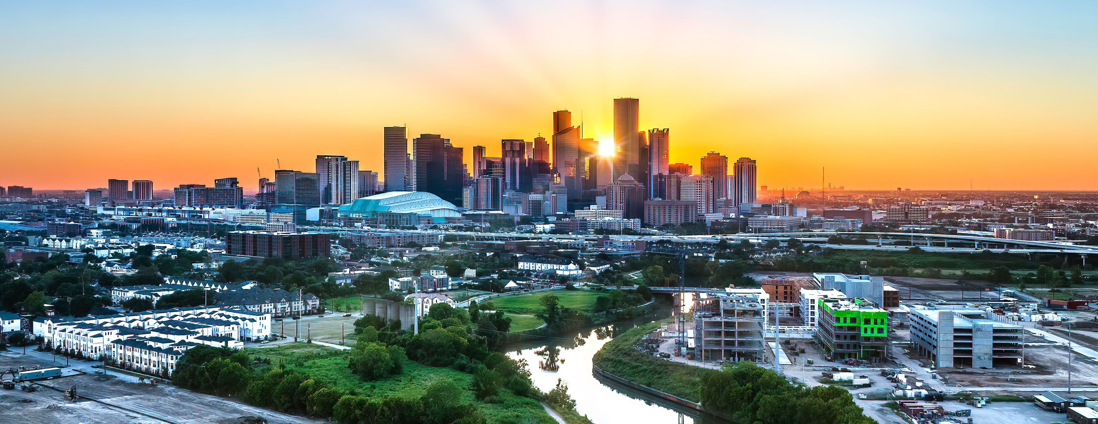A city skyline at sunset with a river in the foreground and a stadium in the center.