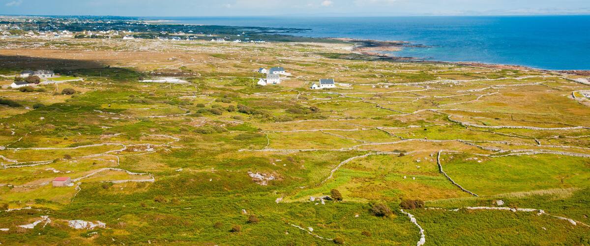 An aerial view of a coastal landscape with stone walls, grassy fields, and a white building near the shore.