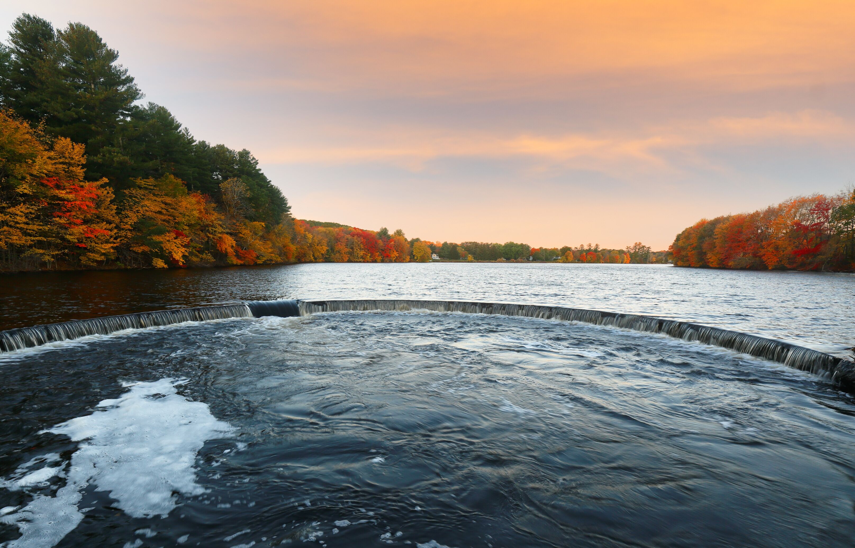 A river with a waterfall, surrounded by autumn-colored trees.