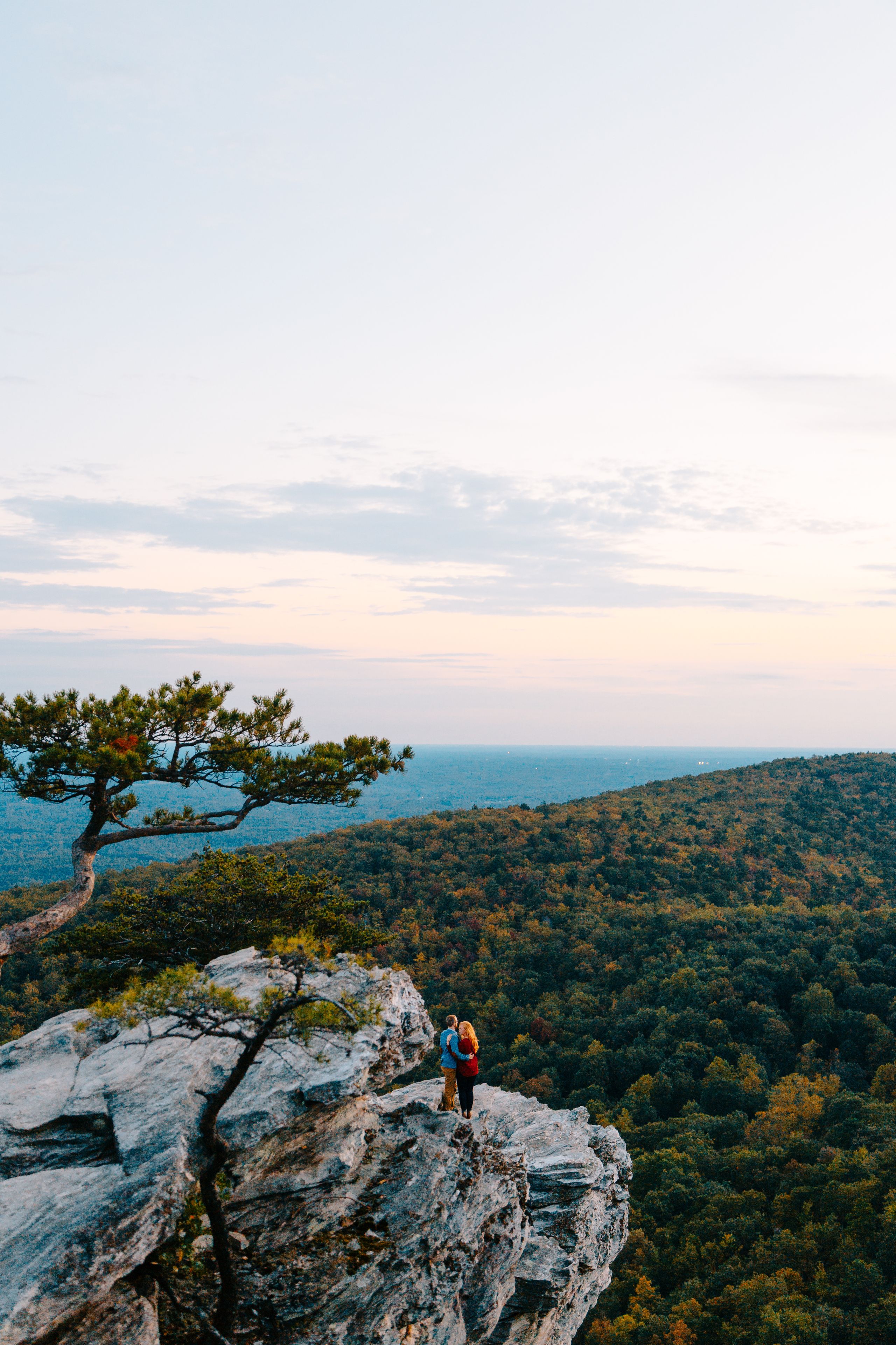A couple standing on a rocky outcrop overlooking a dense forest and a distant body of water.