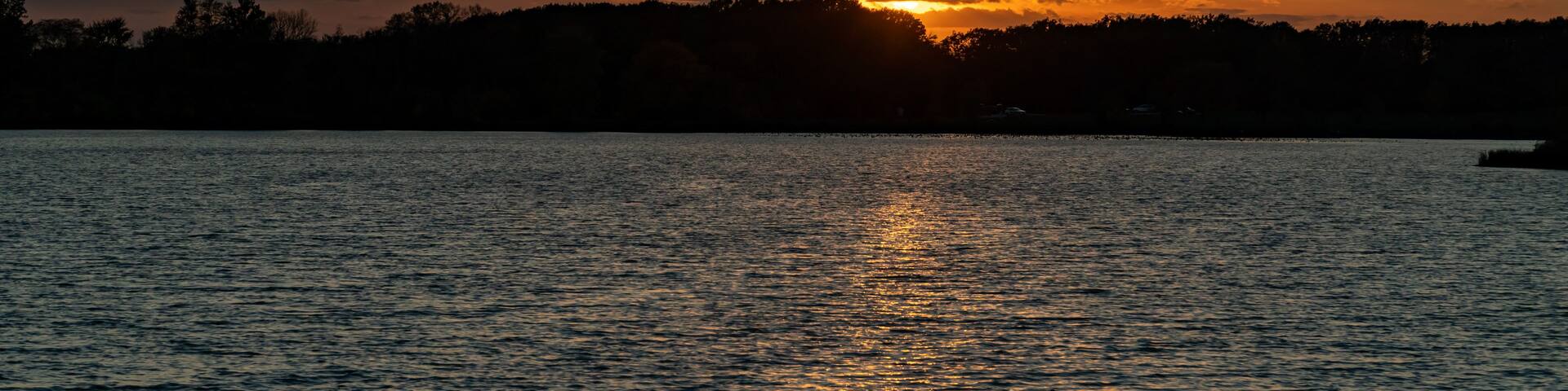 A sunset over a calm lake with trees silhouetted against the sky.